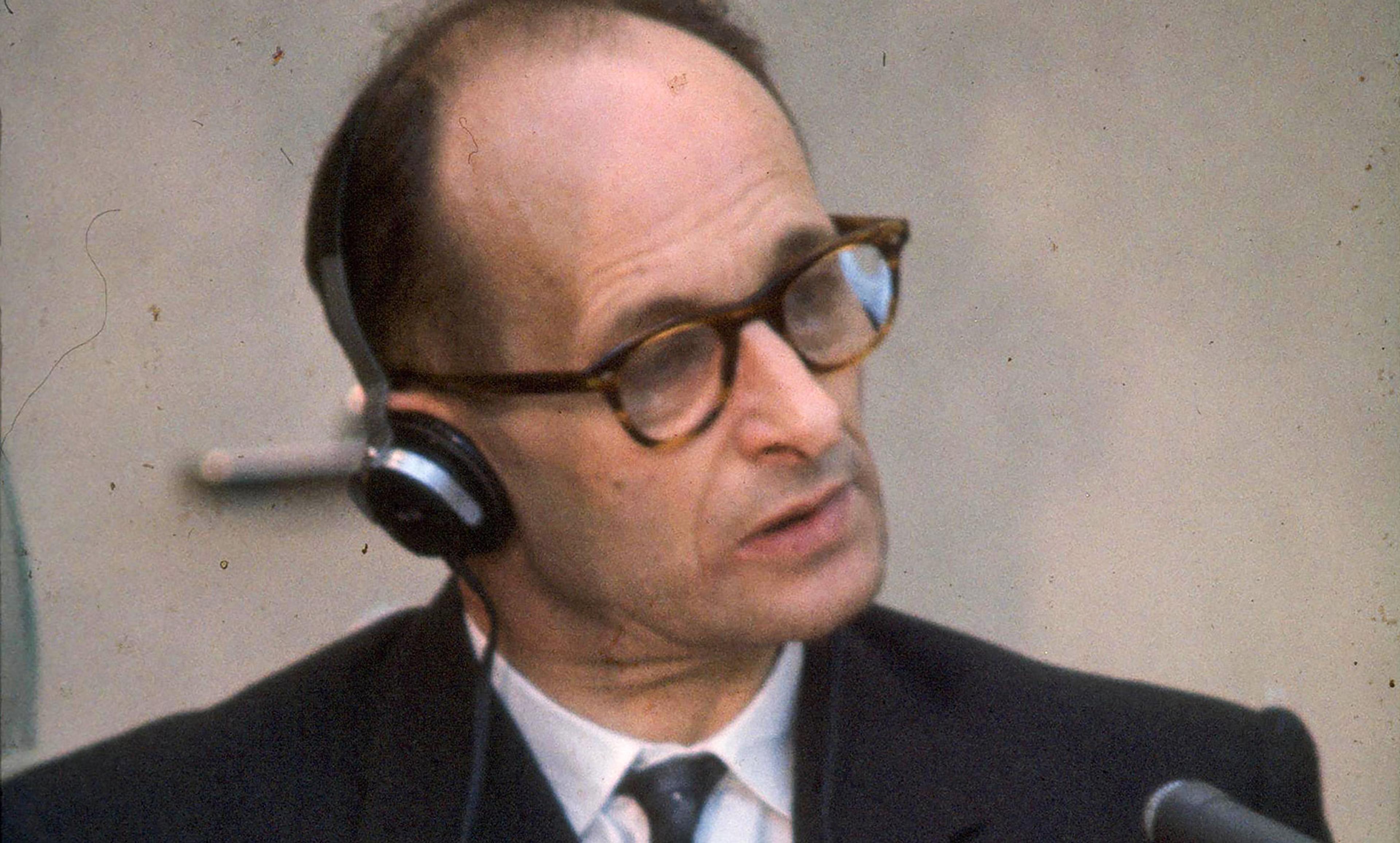 Adolf Eichmann in court with headphones, wearing a suit and tie, looking to the side, against a plain background.