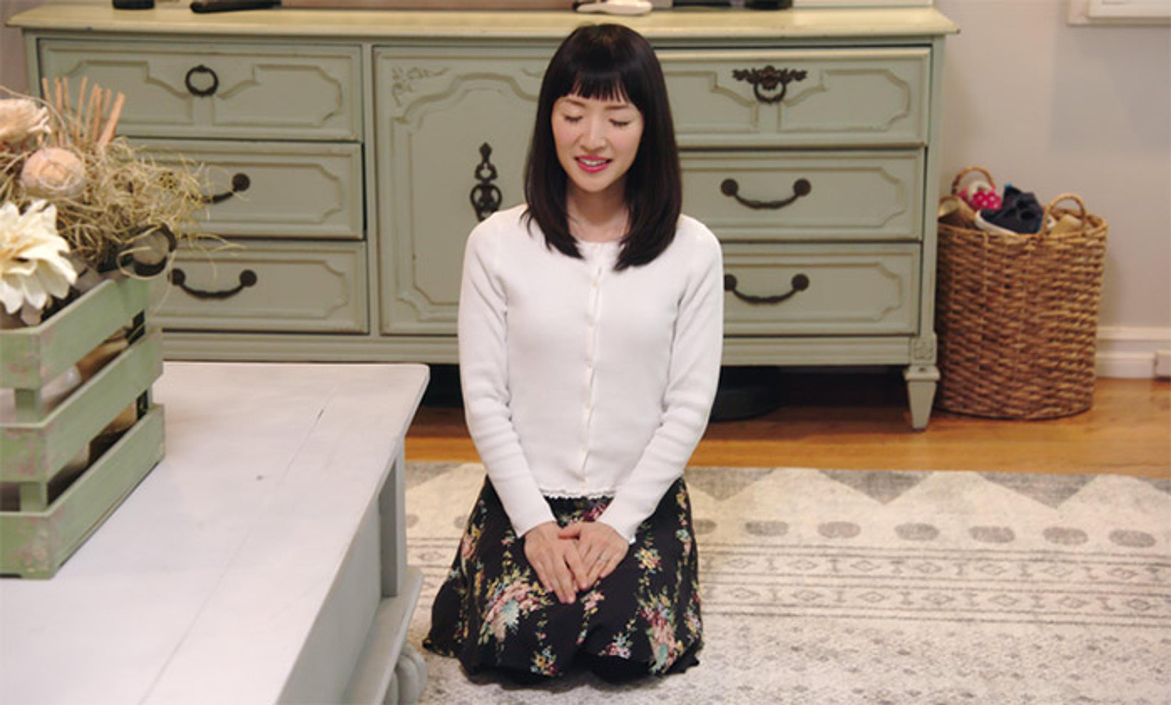 Marie Kondo kneeling with her eyes closed on a patterned rug with hands clasped, wearing a white cardigan and floral skirt in front of a vintage dresser.