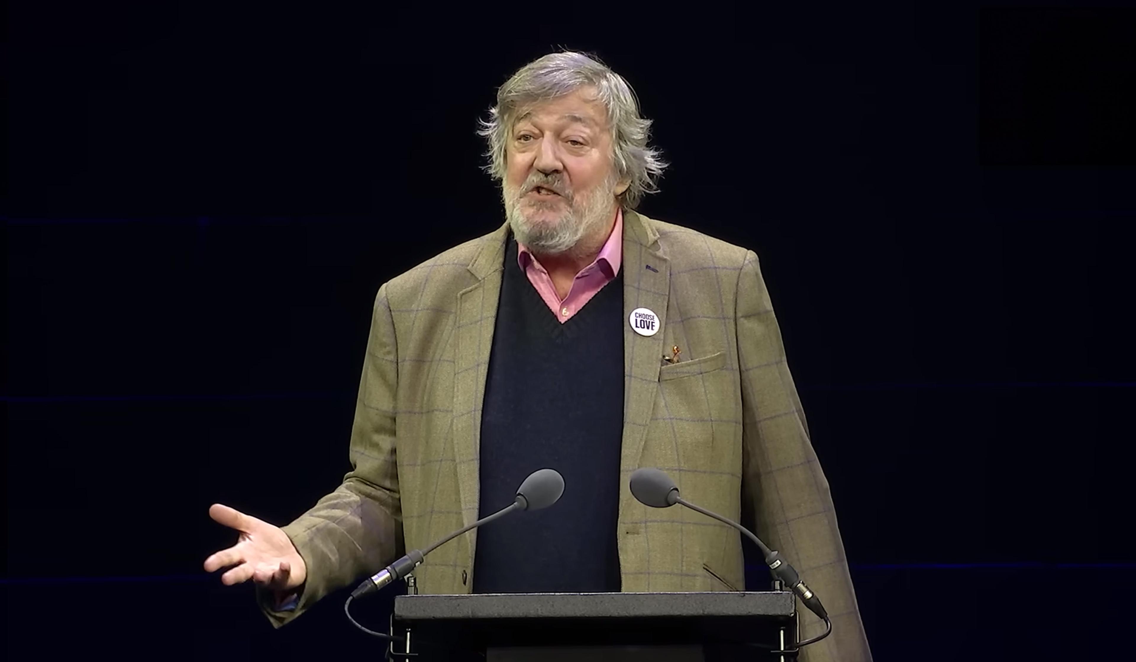 Stephen Fry stands at a podium, wearing a checkered blazer, pink shirt, and ‘Choose Love’ badge, speaking with an expressive gesture.