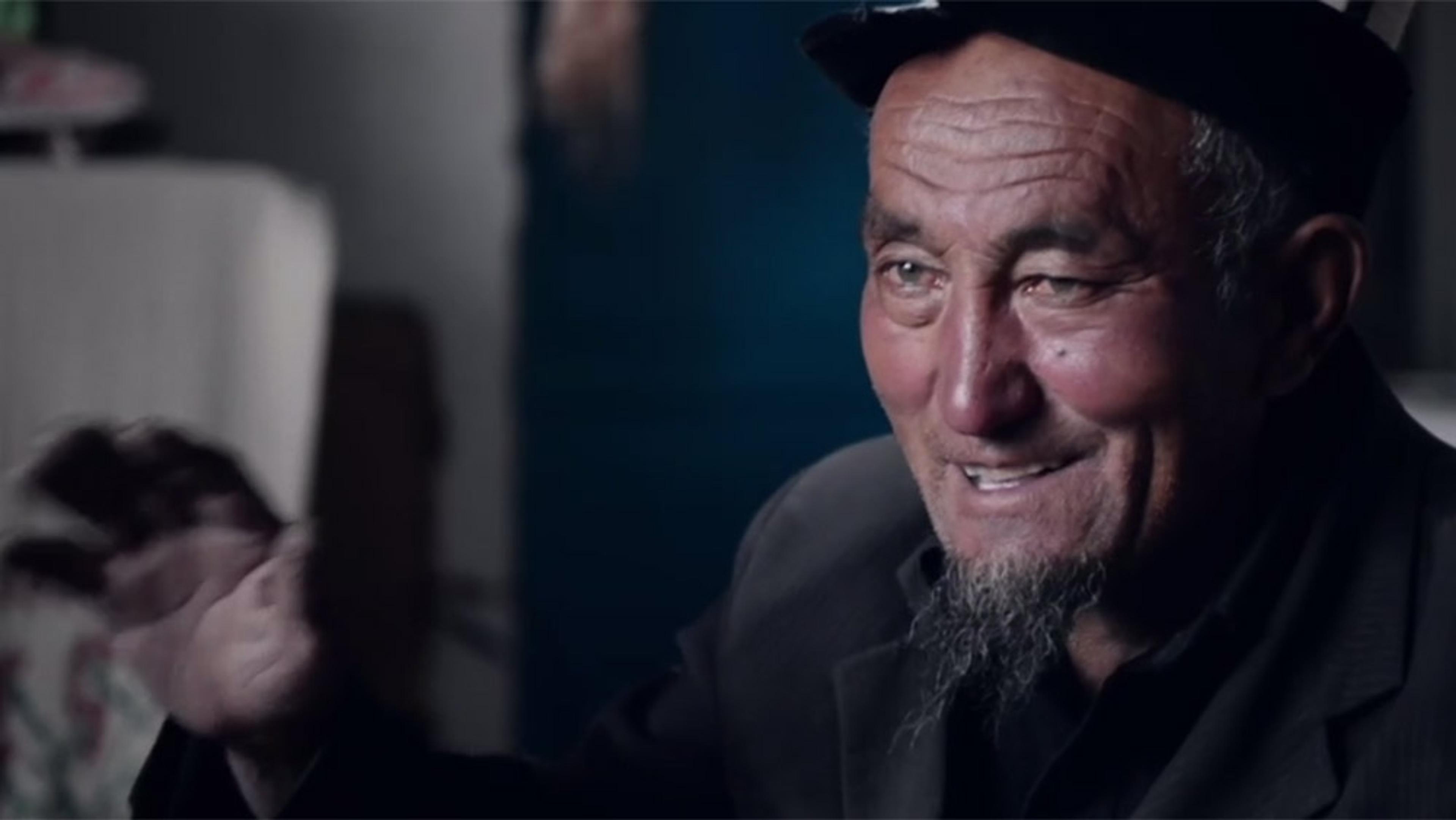 An older man with a beard and cap, smiling warmly with an outstretched hand in the foreground, indoors.