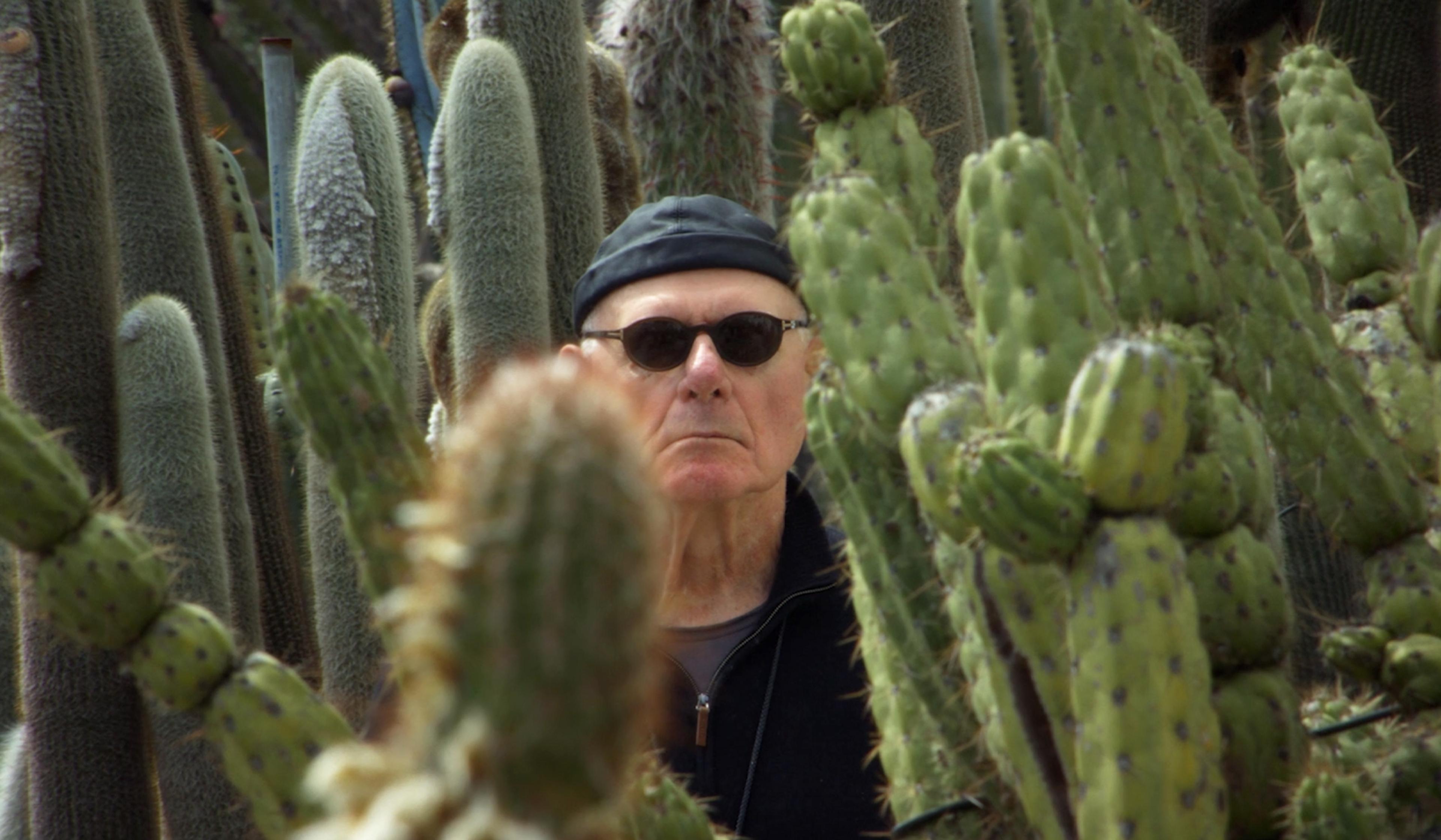 German man wearing sunglasses and a black cap standing among tall, green cacti, with a serious expression on his face.