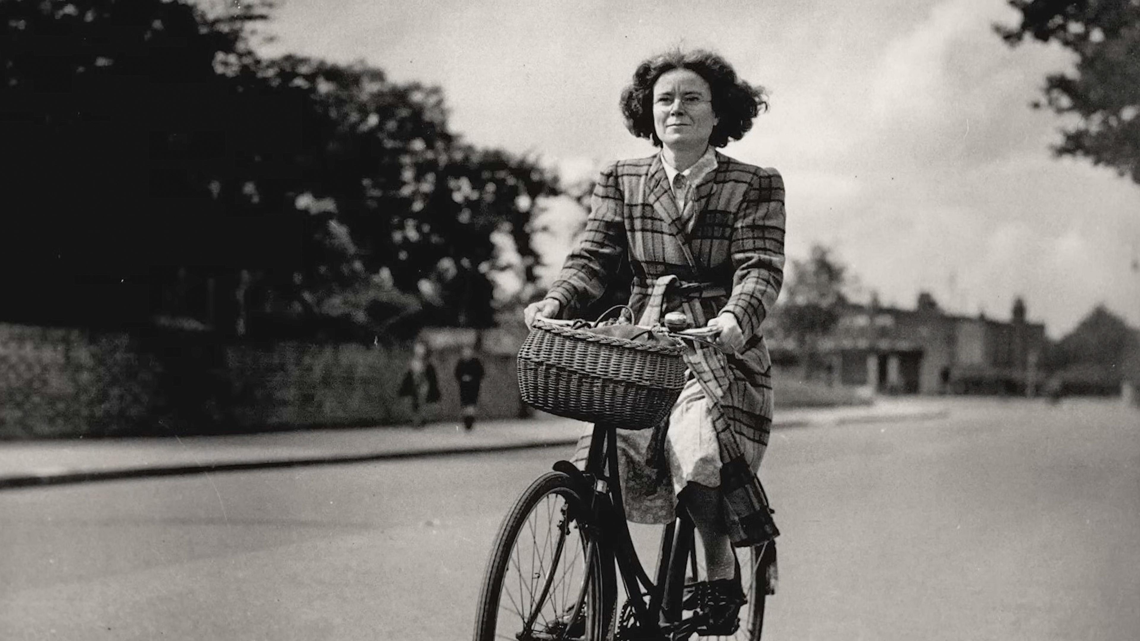 Black and white photo of a woman cycling down a street with a wicker basket on the bike. She wears a checked coat.