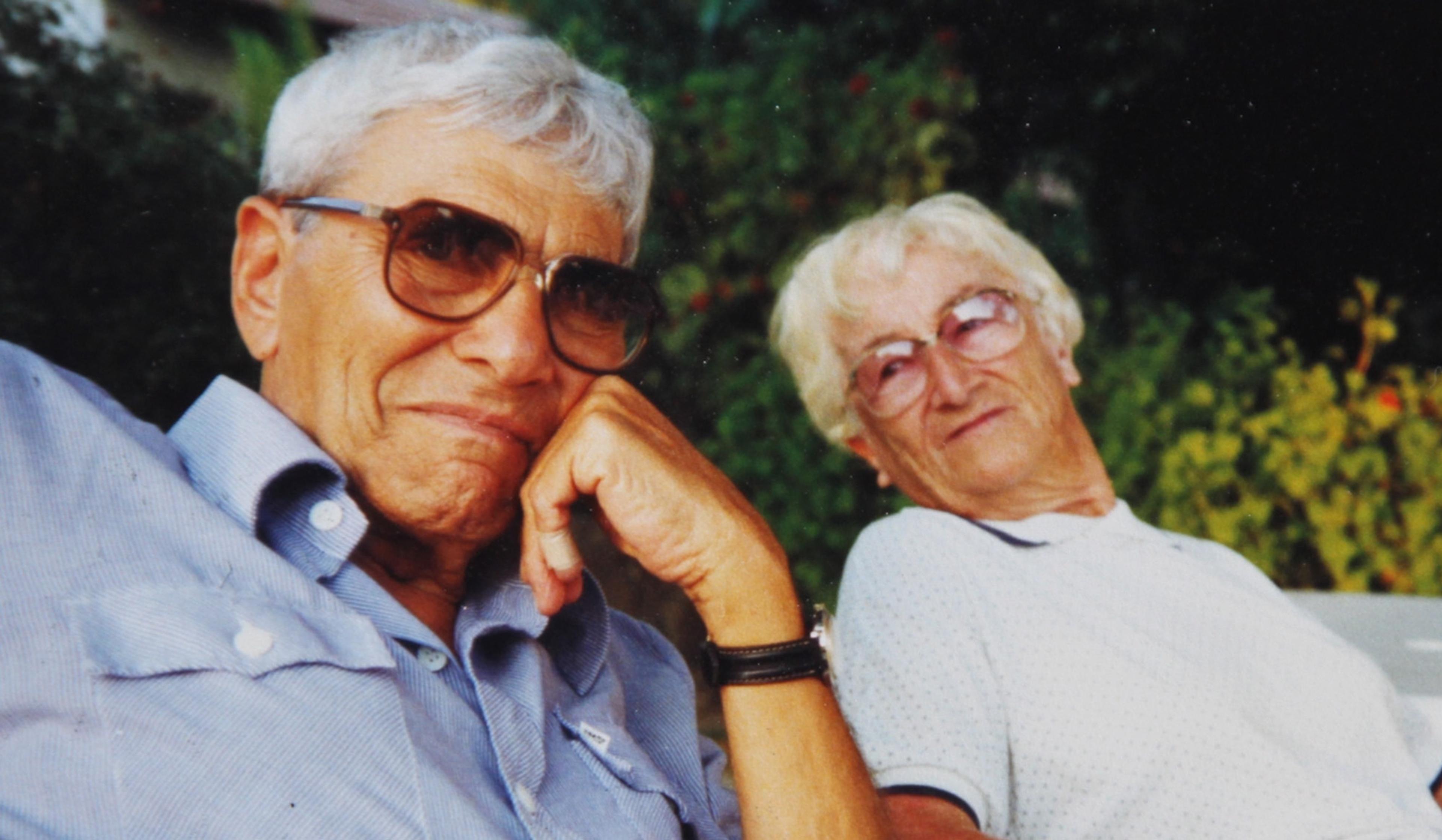 Elderly couple sitting outdoors, both wearing glasses and light-coloured shirts, with greenery in the background.