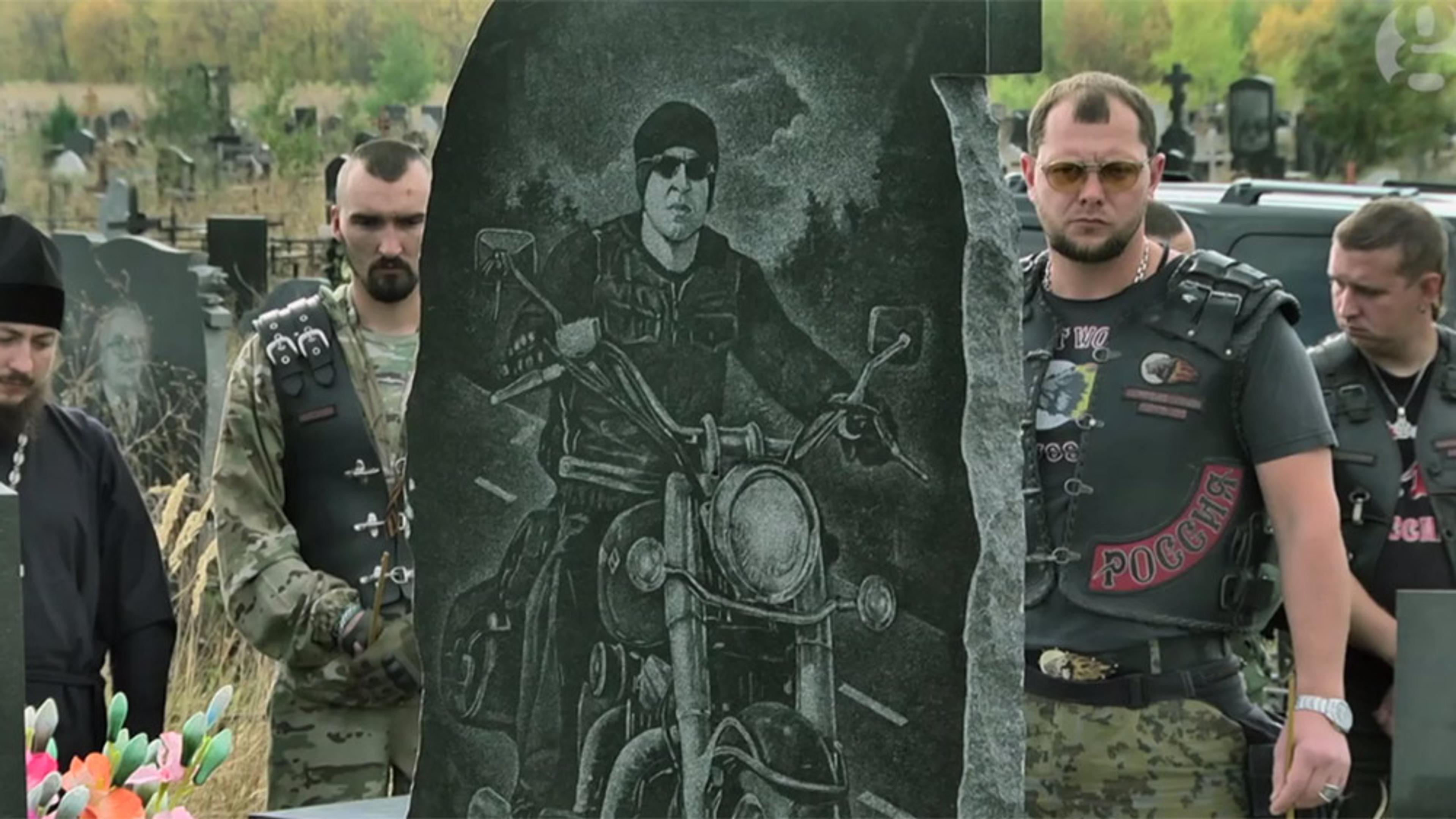 Several men standing around a gravestone in a cemetery. The gravestone features an engraving of a biker in sunglasses.
