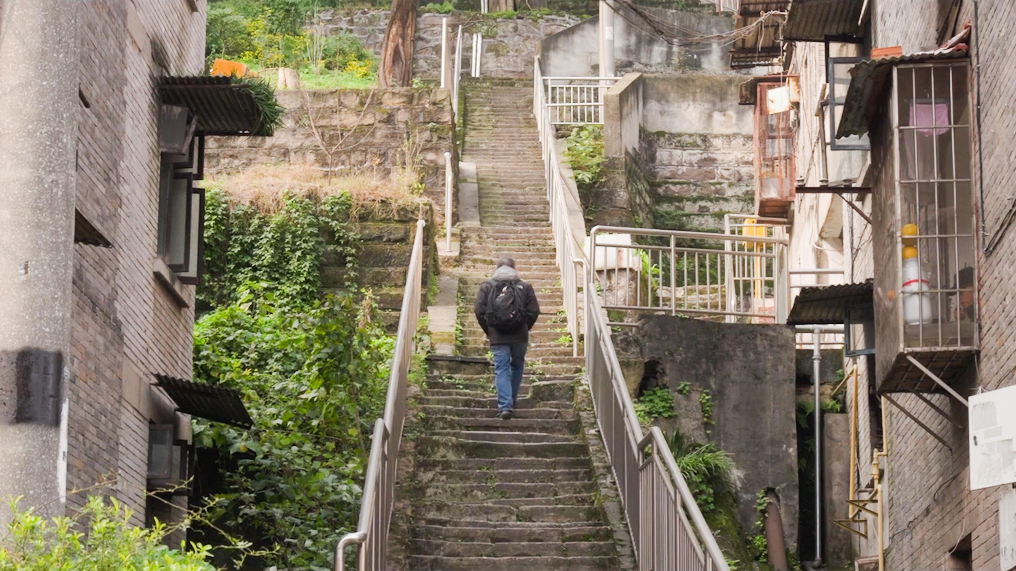 Photo of a person with a backpack walking up a narrow outdoor staircase between brick buildings and lush greenery.